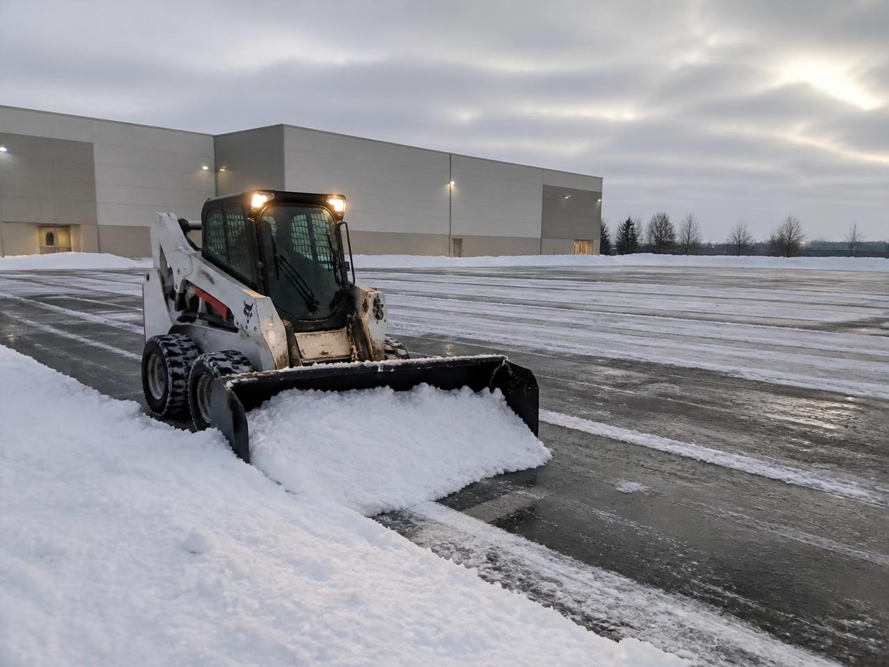 Skid steer with a snow pusher box clearing a commercial parking lot at dawn in Ocean County, New Jersey